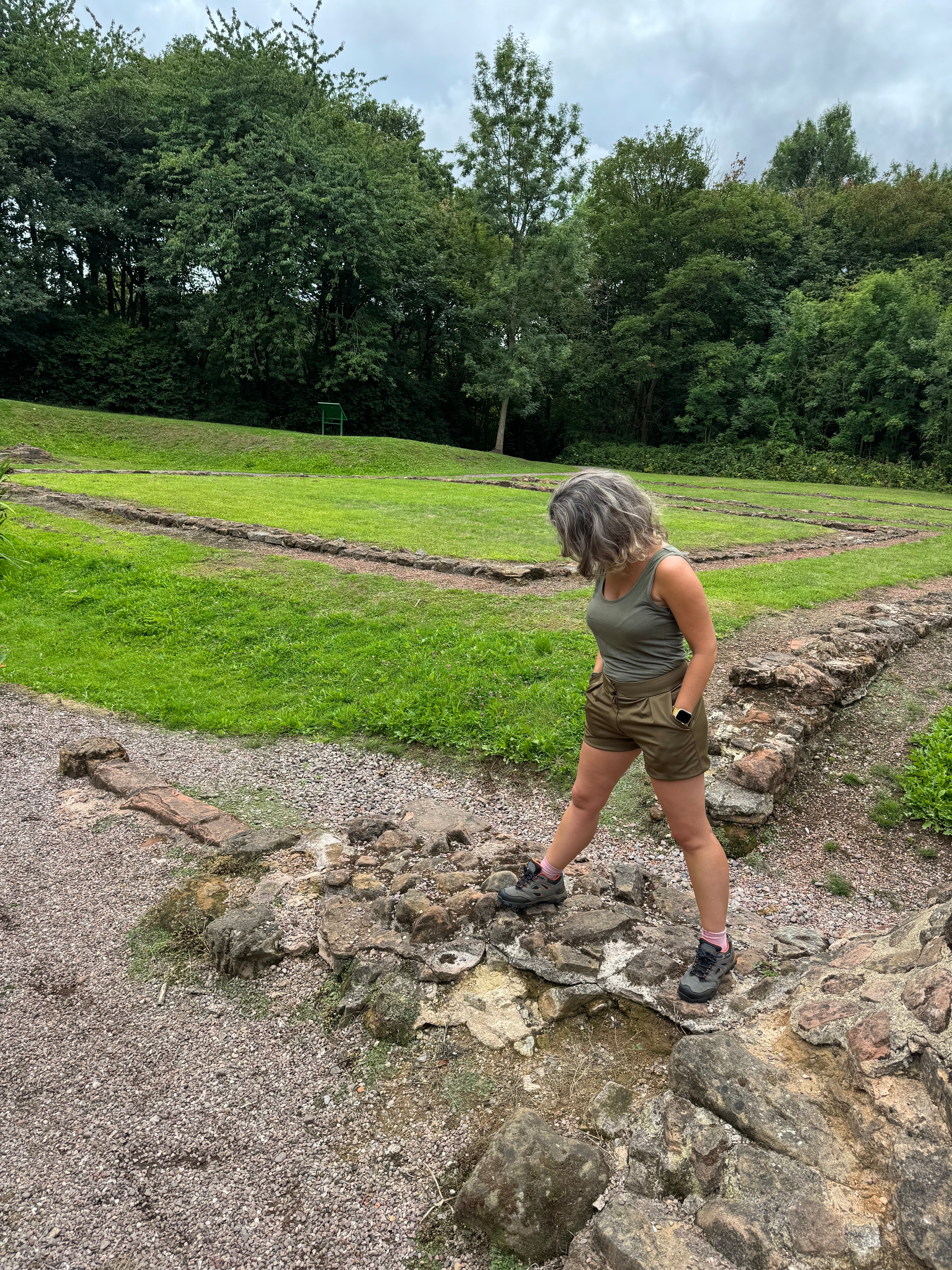 Su Dias, founder of P’tit Affair, wearing olive green petite tailored shorts while standing on the ruins at Sandwell Priory, surrounded by greenery