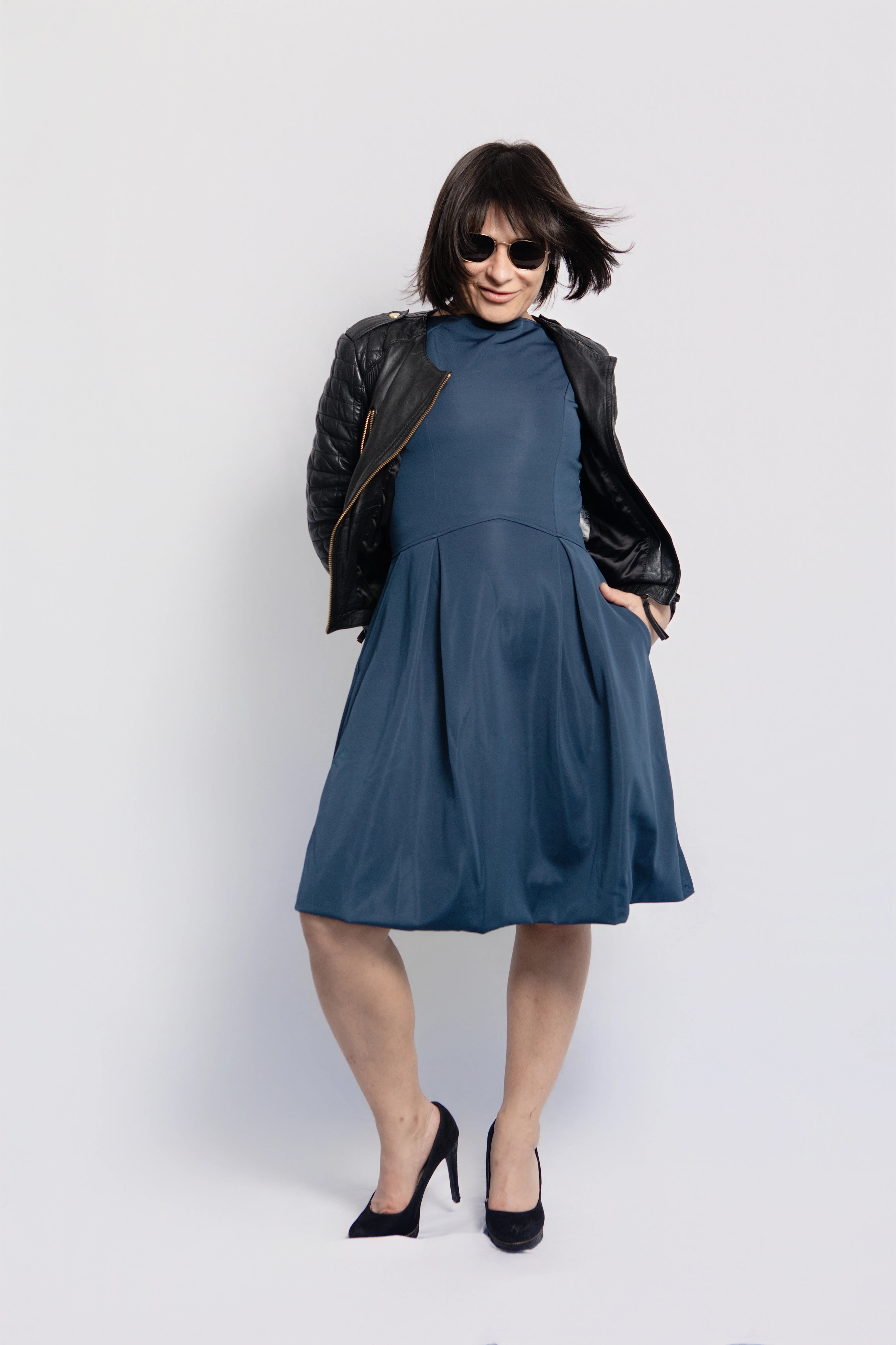 Petite woman wearing a blue midi dress with pockets styled with a black leather jacket and heels, showing a versatile day-to-night look against a white background