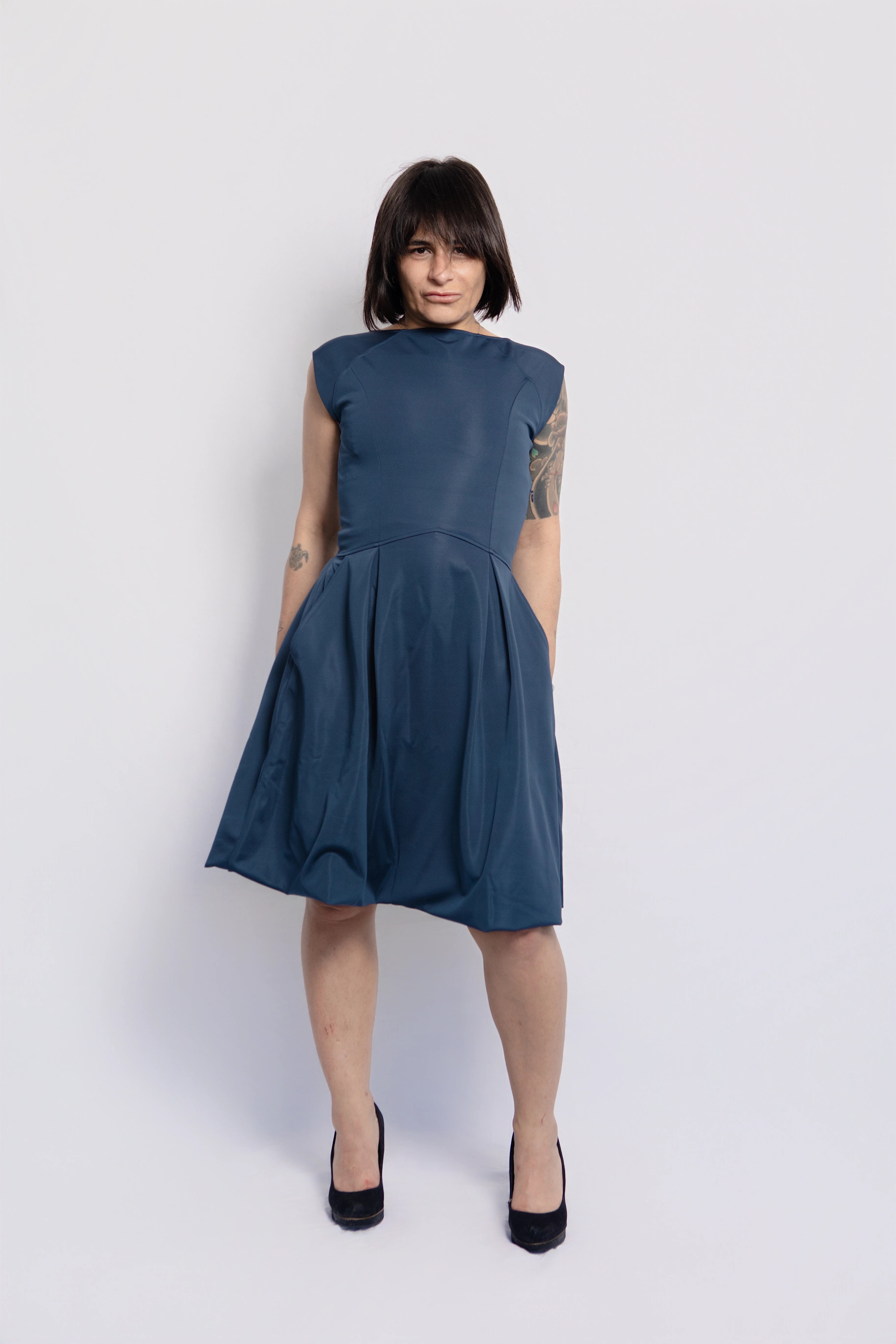 Full-length view of a woman wearing a blue petite midi dress with pockets, designed for women 5'3 and under, styled with black heels against a white background