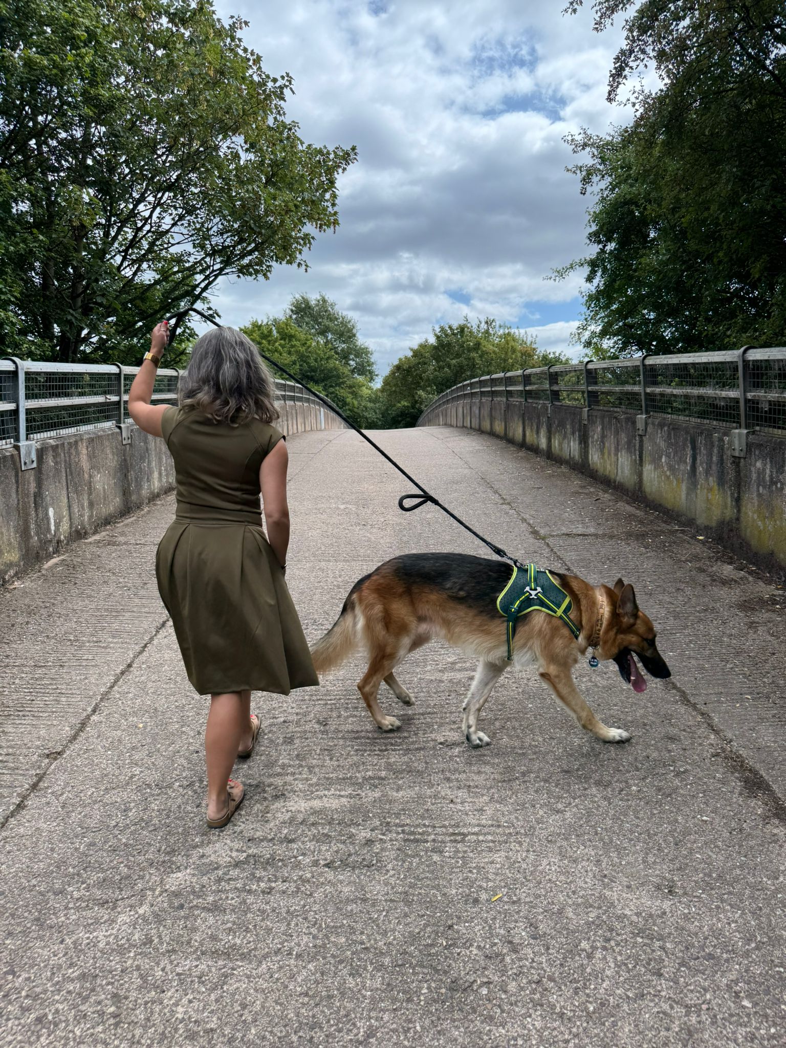 Petite woman wearing P’tit Affair Eloise Midi Dress in olive, walking her German Shepherd across a bridge outdoors