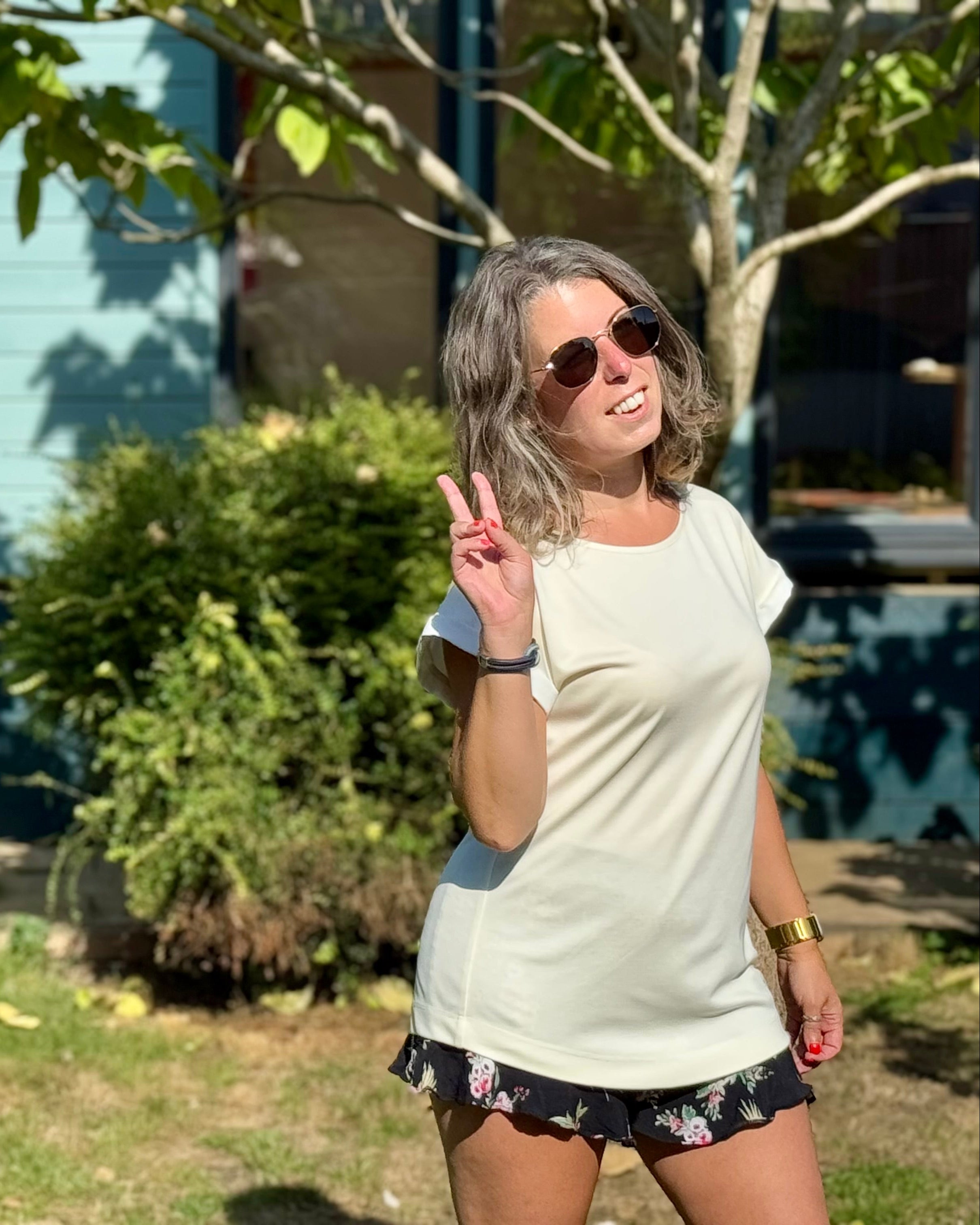 Petite woman wearing the P’tit Affair Millie T-shirt in white, styled casually outdoors with sunglasses and floral shorts.