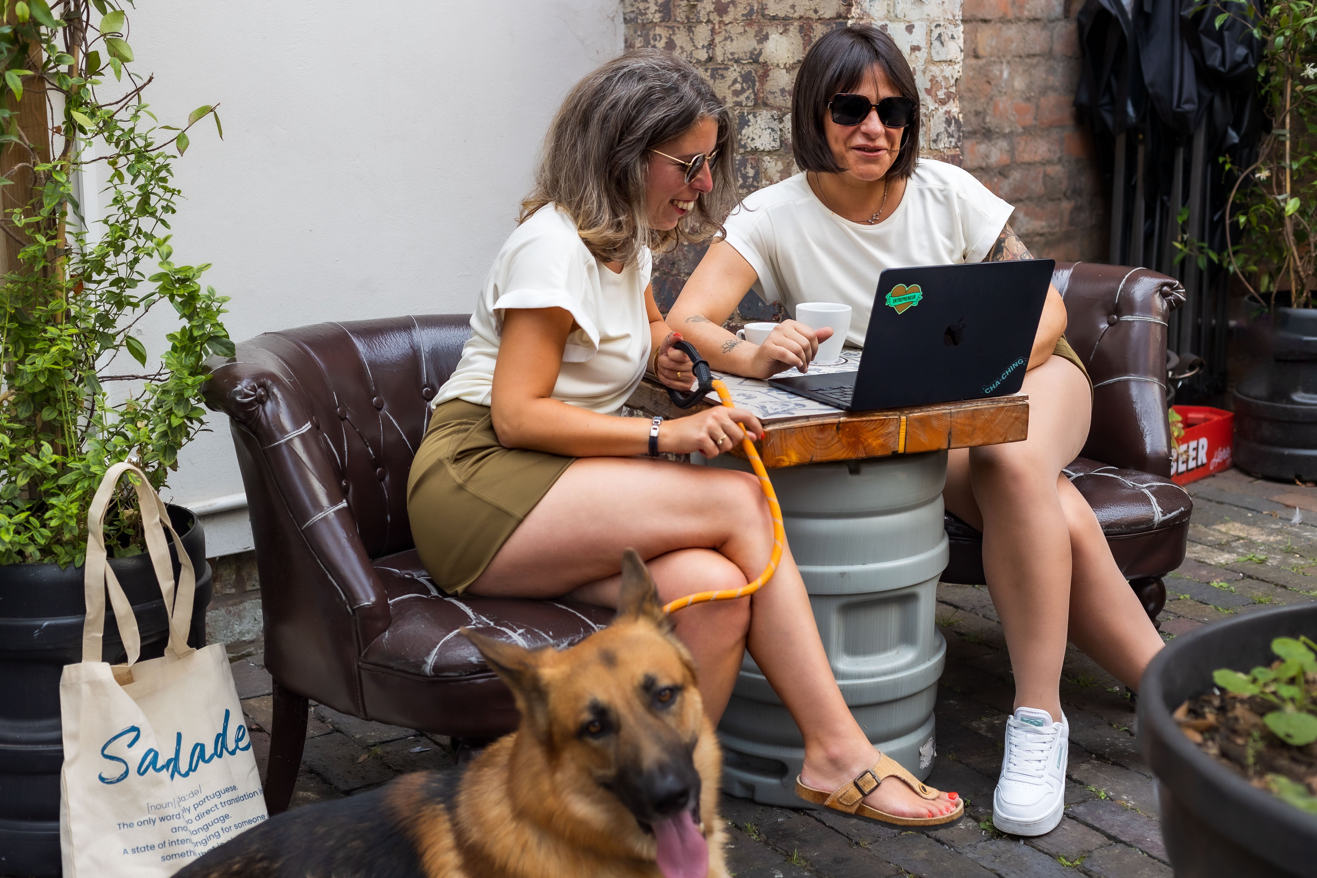 Two petite women sitting outdoors working on a laptop, wearing P’tit Affair pieces, with a foldable Saudade Tote Bag and a German Shepherd nearby