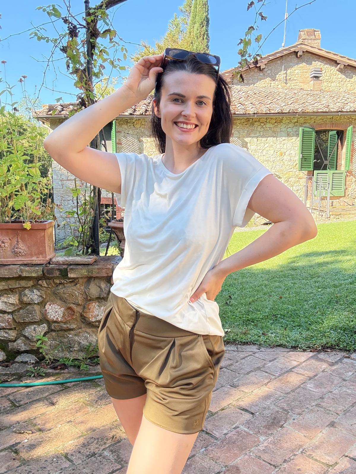 Model wearing P’tit Affair’s Millie T-shirt in Vapourous Grey and Mariana Tailored Shorts in Olive Green, smiling outdoors. Timeless capsule wardrobe designed for petite women 5'3" and under.